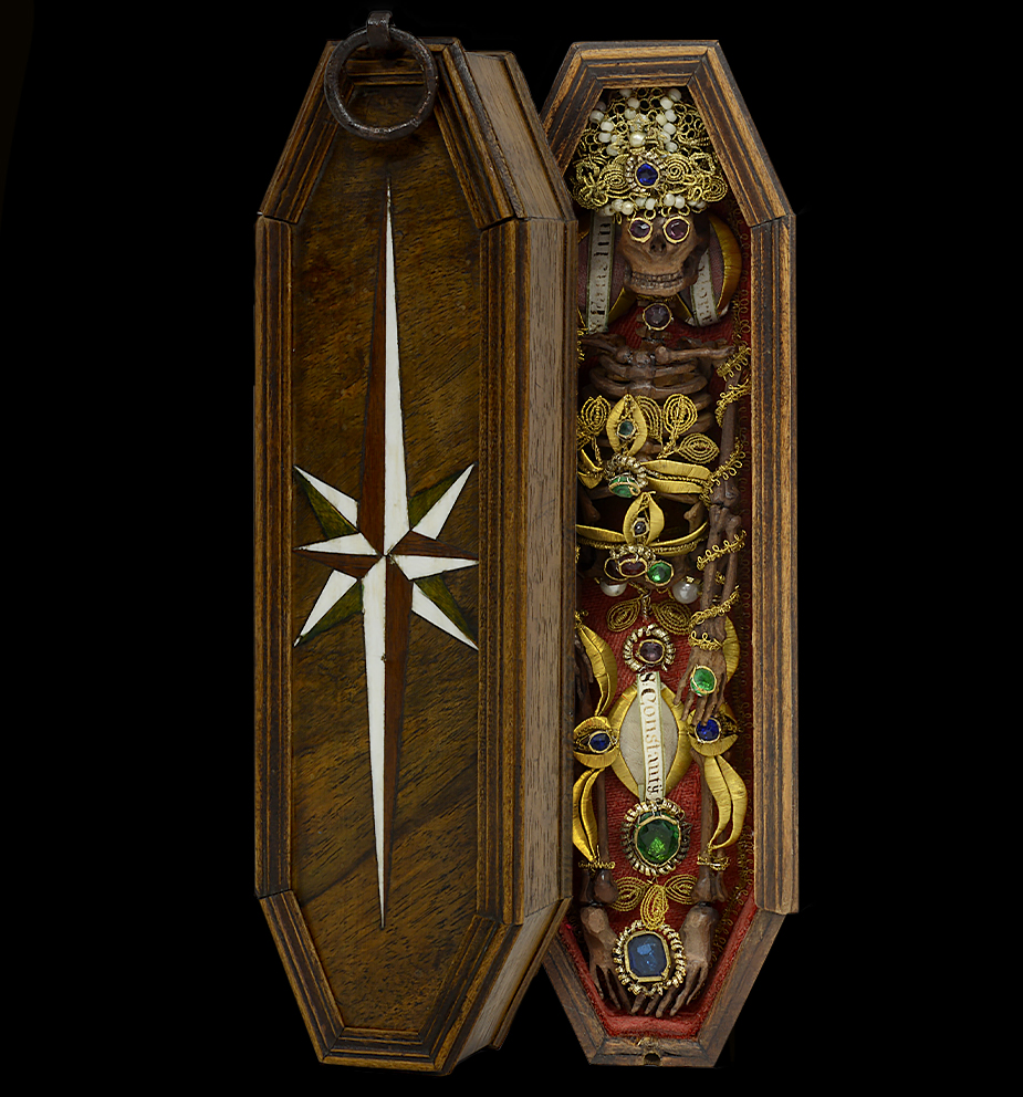 Memento Mori, Süddeutsch Siehe auch Rubrik »Objekt des Monats«. Memento Mori in der Art der Hl. Leiber. Das anatomisch korrekte Skelett aus Birnbaumholz geschnitzt. Die Verzierungen in Silber und Golddraht, die Steine aus buntem Glas. Der innenliegende rote Stoff ein Wolldamast. Die Reliquien mit gemalten Pergament Cedulae. Der Sarg in Nussbaumholz mit Profilleisten. Die Sternintarsie auf dem Schiebedeckel aus Bein, Mahagoni und grün gefärbtem Ahorn. Der Verschluß ein Eisenring mit Gewinde. Maße: L 23 X B 6,5 X H 4 cm Süddeutsch, Bayern Barock, Mitte 18. Jahrhundert