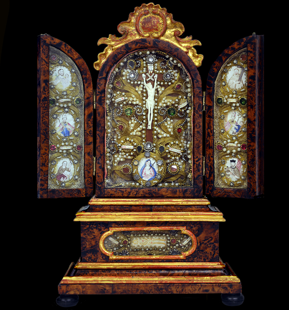 Klappaltar, Süddeutsch Im Mittelfeld, gekreuzigter Christus in Bein. Am Fuß des Kreuzes die Schmerzhafte Maria in Gouachemalerei auf Pergament. Die Szene mit zahlreichen Reliquien umrandet. Die Klosterarbeiten, Gold und Silberdraht, Perlen und Glassteinen. Die Seitenflügel mit Heiligenabbildungen und Reliquien. In der Predalla, Hl. Tuch von Turin, rückseitig gesiegelt. Das Gehäuse furniert mit Erlenkropfholz. Der Aufsatz und die Leisten original vergoldet. Die Beschlagteile Messing graviert. Maße: geschlossen: H 40 x B 23 x T 10 cm, geöffnet: H 40 x B 30 x T 10 cm Bayern 1. Hälfte 18. Jahrhundert